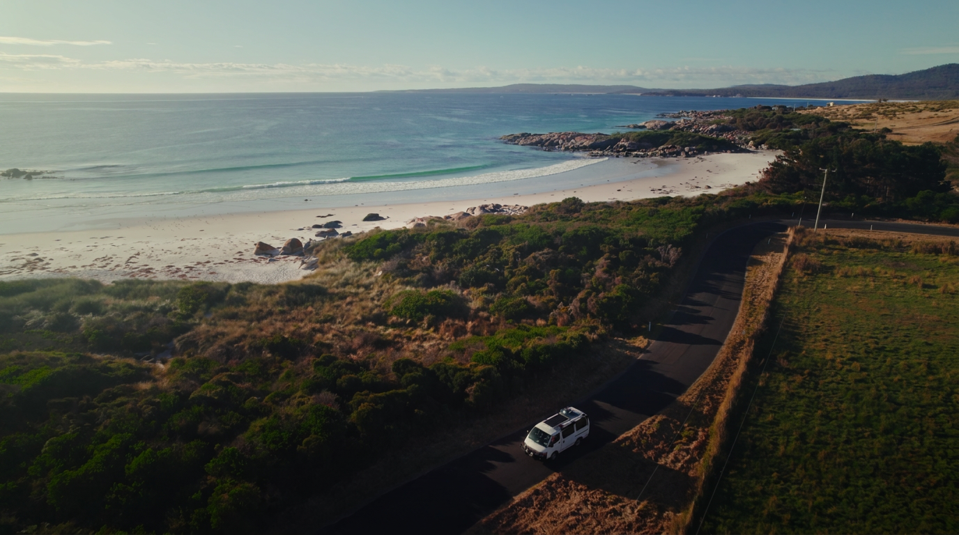 Aerial view of a van driving along a coastal road beside a white sand beach